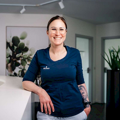 Porträt von Kathrin Raffler, Mitarbeiterin in der Zahnarztpraxis steht lächelnd in blauem Shirt vor Praxisempfang.