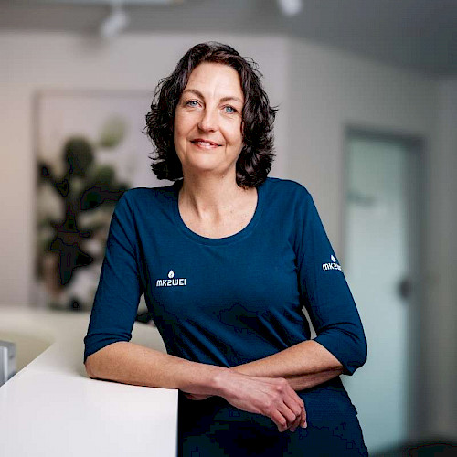 Porträt von Andrea Gropper, Mitarbeiterin in der Zahnarztpraxis steht lächelnd in blauem Shirt vor Praxisempfang.