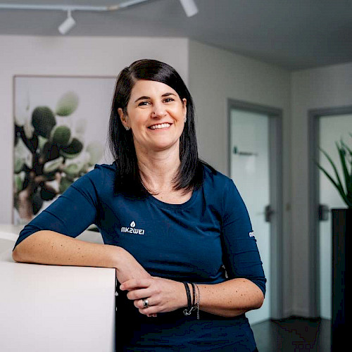 Porträt von Kerstin Rock, Mitarbeiterin in der Zahnarztpraxis steht lächelnd in blauem Shirt vor Praxisempfang.